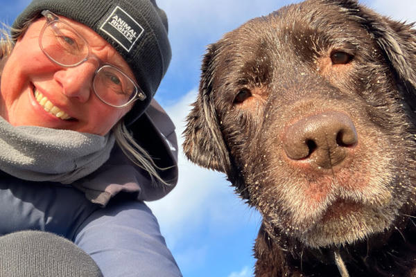Linda met jas en muts op en haar bruine labrador Brownie die samen recht in de camera kijken, met de blauwe buitenlucht op de achtergrond.